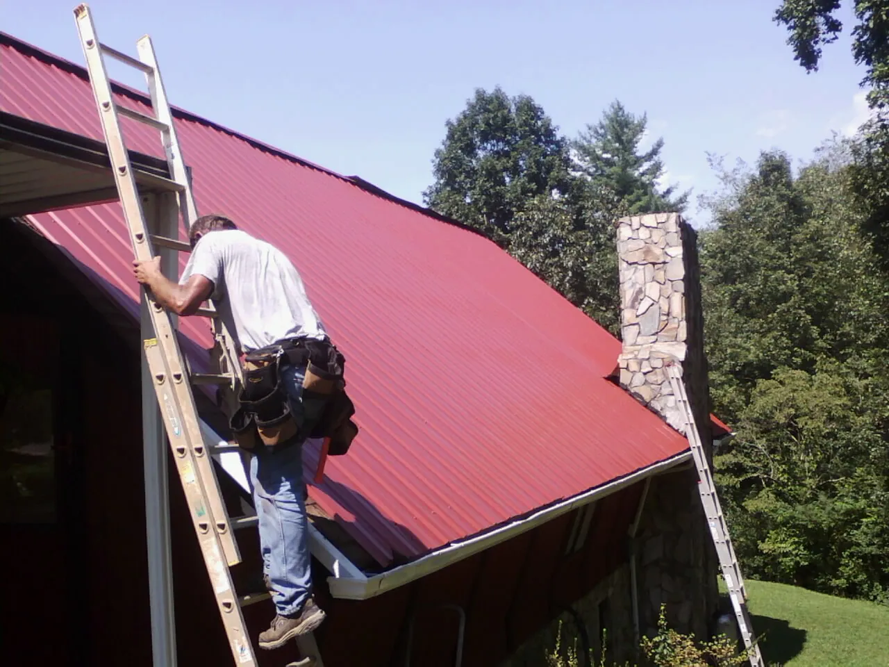 Roofer on a ladder beside a red metal roof and stone chimney — Soffit & Fascia Repair by Roofing Brothers in Cicero
